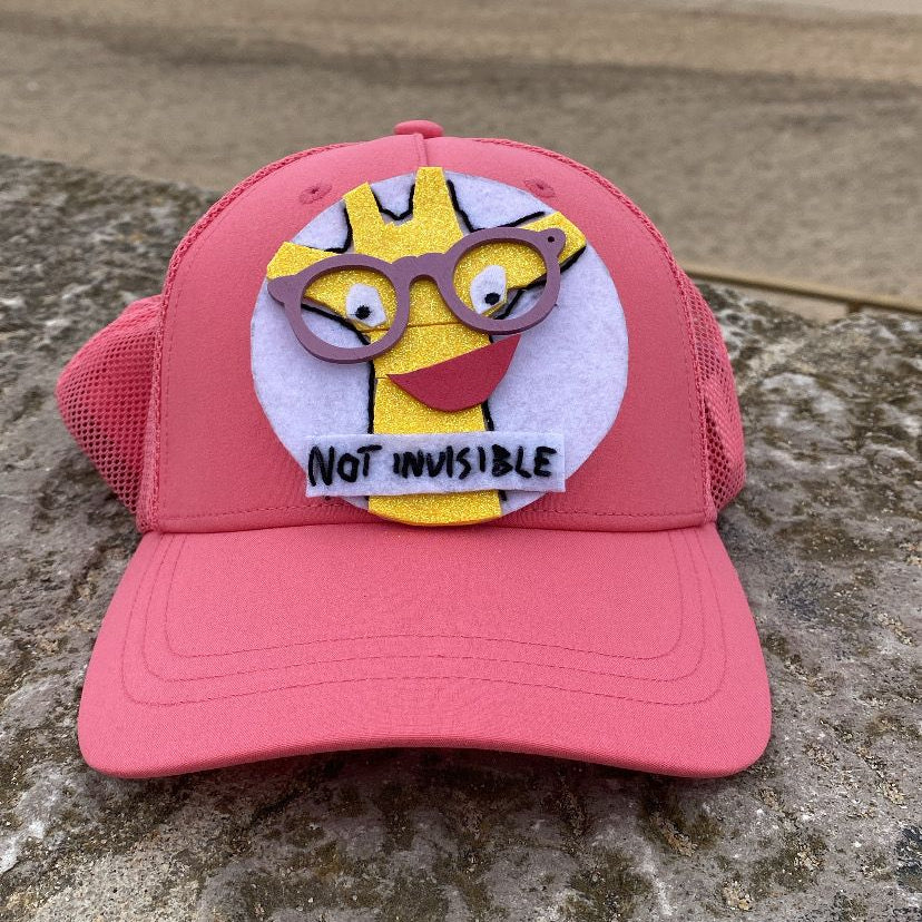 Pink cap with cartoon character and 'Not Invisible' text on a stone surface with beach in the background